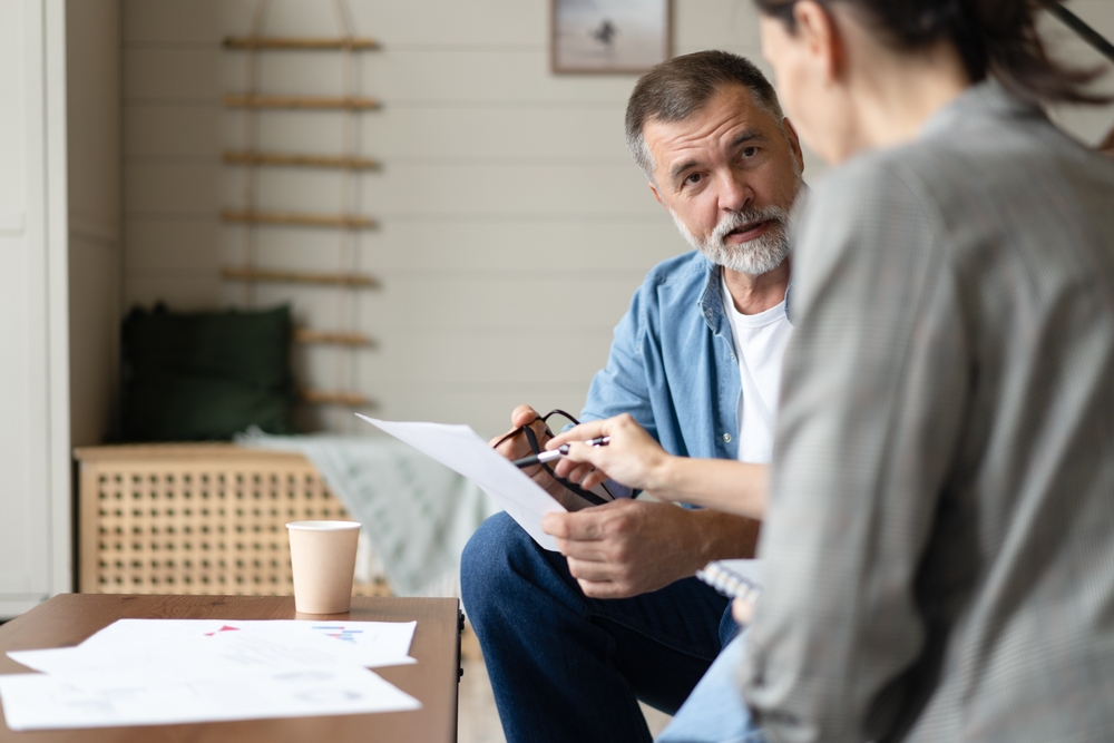 Older bearded man in a blue shirt discussing a printed document with an adviser across a coffee table, representing a retirement planning conversation for a Lake Macquarie client