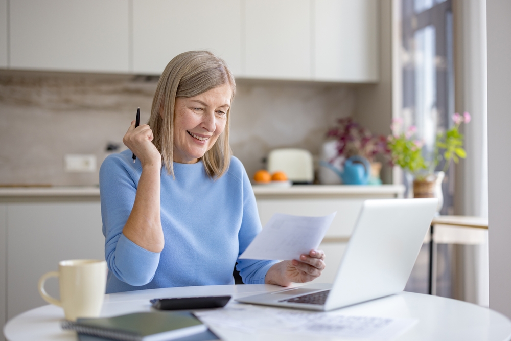 Smiling older woman in a blue jumper reading her retirement plan paperwork at a kitchen table with a laptop and calculator, representing retirement planning for a Port Stephens client