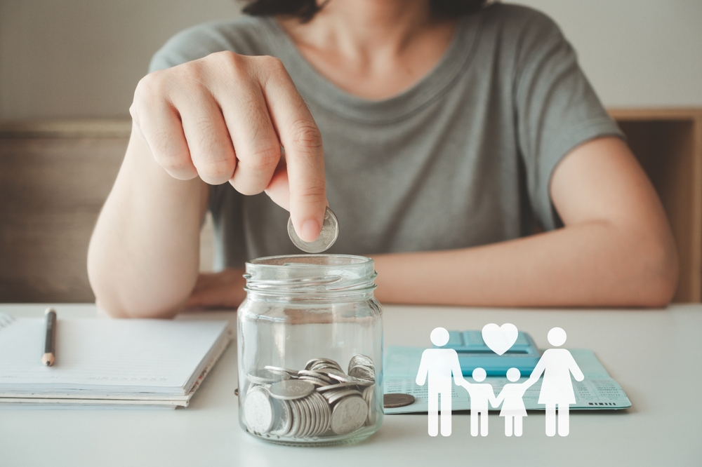 Woman dropping a coin into a glass jar of savings beside a notebook and a paper family cut-out, representing superannuation contributions for a Lake Macquarie client