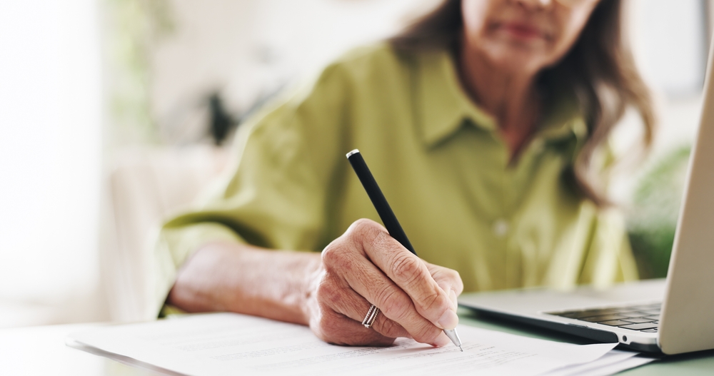 Close-up of an older woman in a green shirt writing notes on a retirement planning document beside a laptop at home, representing retirement planning for a Hunter Valley client