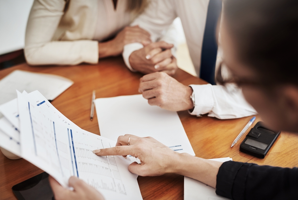Three people seated around a wooden desk reviewing printed financial charts and notes during a financial planning session for a Port Stephens client of Newburn Wealth Advisory