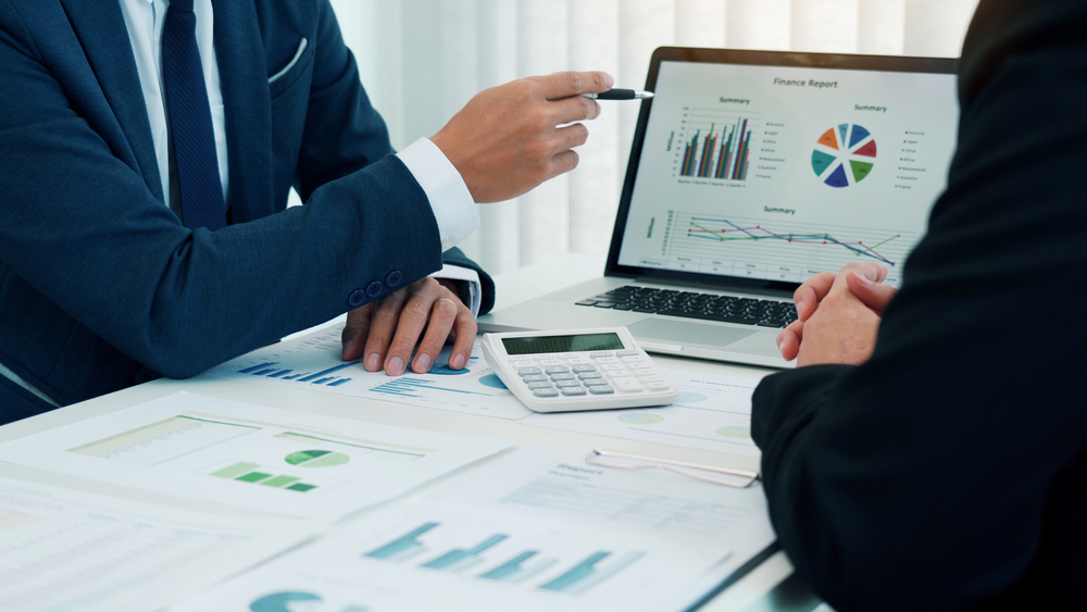 Adviser in a suit pointing at a laptop screen showing finance charts beside a calculator and printed reports during a household financial planning session for a Hunter Valley client